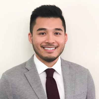 The image features a person standing in front of a plain background. They are wearing a light grey blazer over a dark shirt, paired with a maroon tie, and have short hair. The individual is smiling and appears to be posing for the photo.