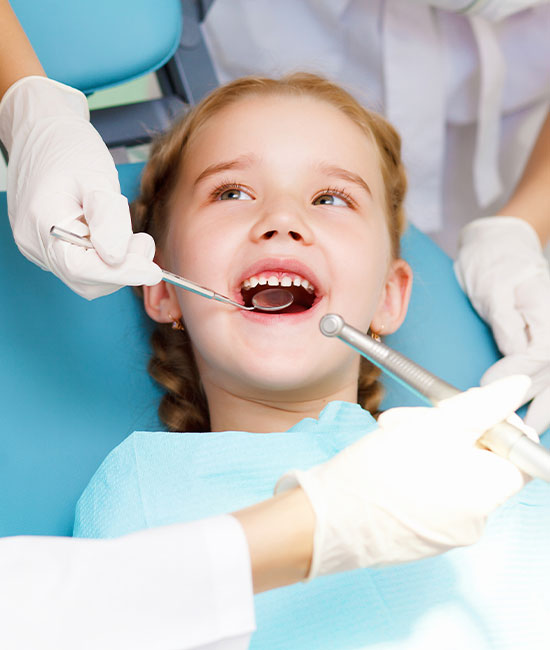 A young girl with a big smile is seated in a dental chair, receiving dental care, with a dentist and dental hygienist attending to her.