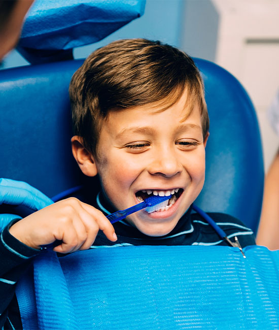 The image shows a young boy sitting in a dental chair with his mouth open, wearing a blue bib, and having his teeth cleaned by a dental professional who is smiling at the camera.