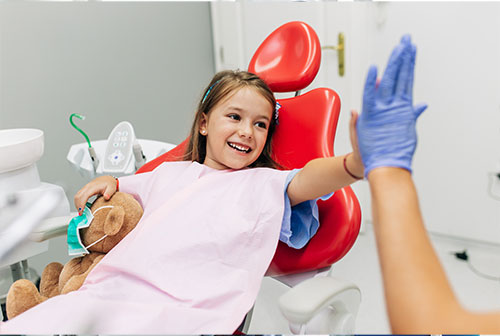 A young girl sitting in a dental chair with a smile on her face, waving at someone off-camera, while a dentist takes a photo of her from behind.