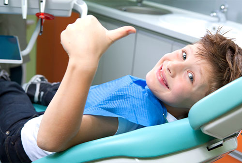 A young boy sitting in a dental chair, smiling at the camera with thumbs up.