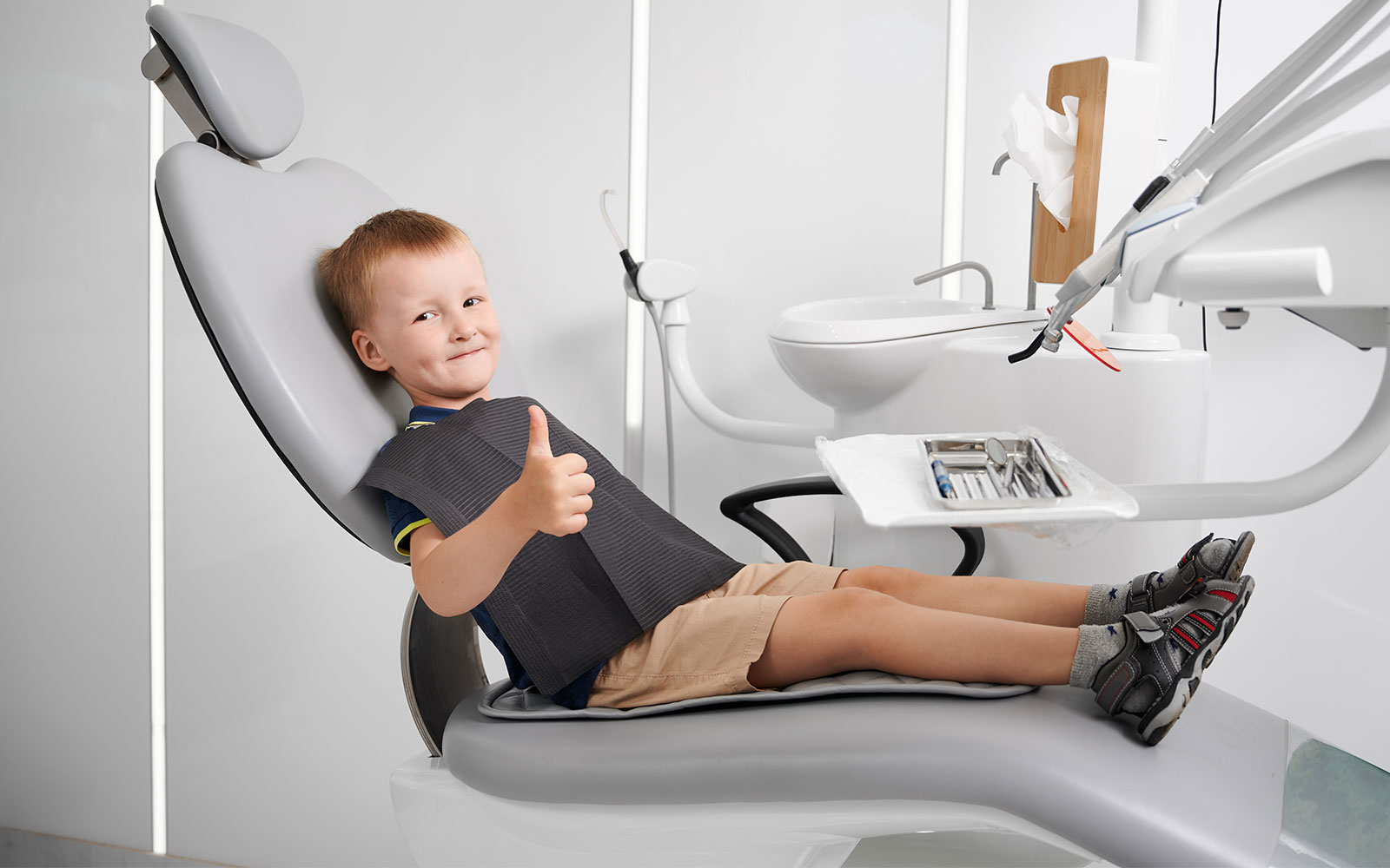 A young boy sitting in a dental chair, smiling at the camera, with his feet on a footrest.