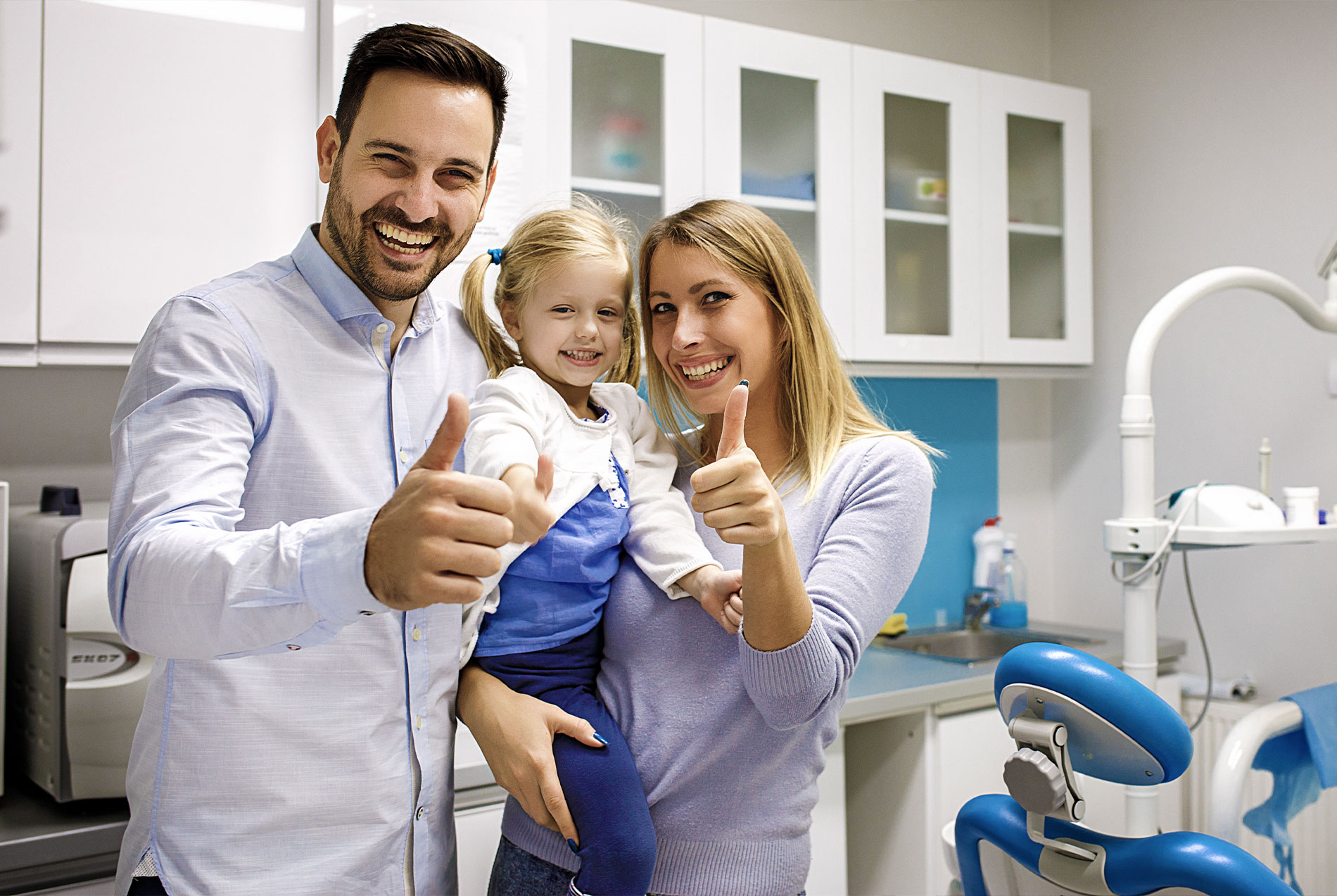 The image shows a family posing together in a dental office setting, with the father giving a thumbs-up gesture.